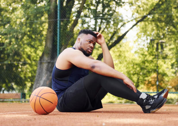 Tired african american man sits on the ground with ball on the court outdoors. Takes a break.